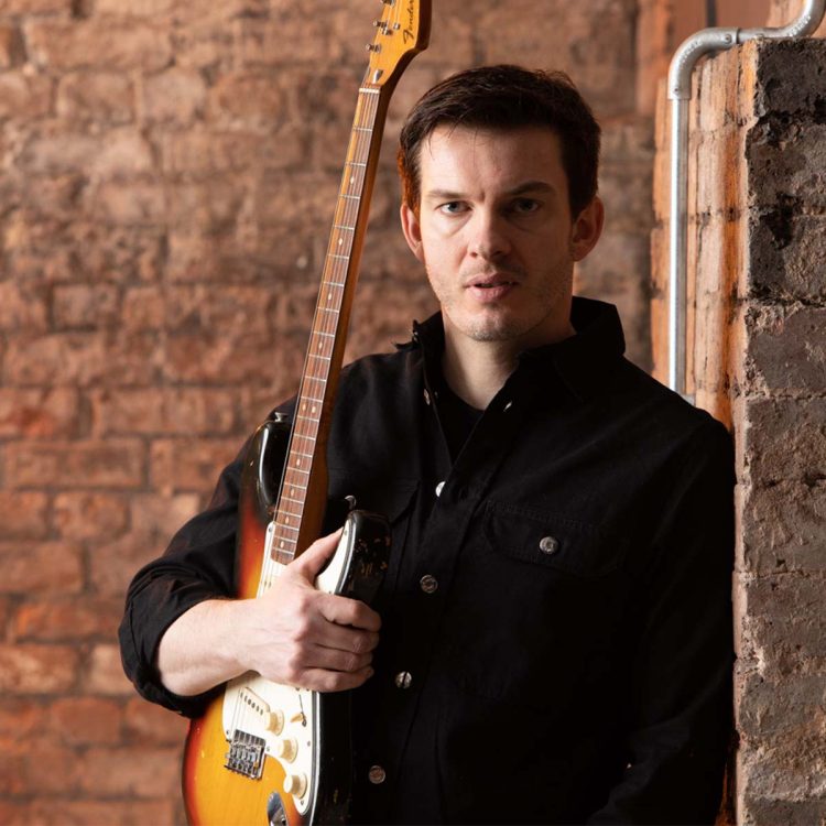 Dan Machin leans against brick wall with guitar