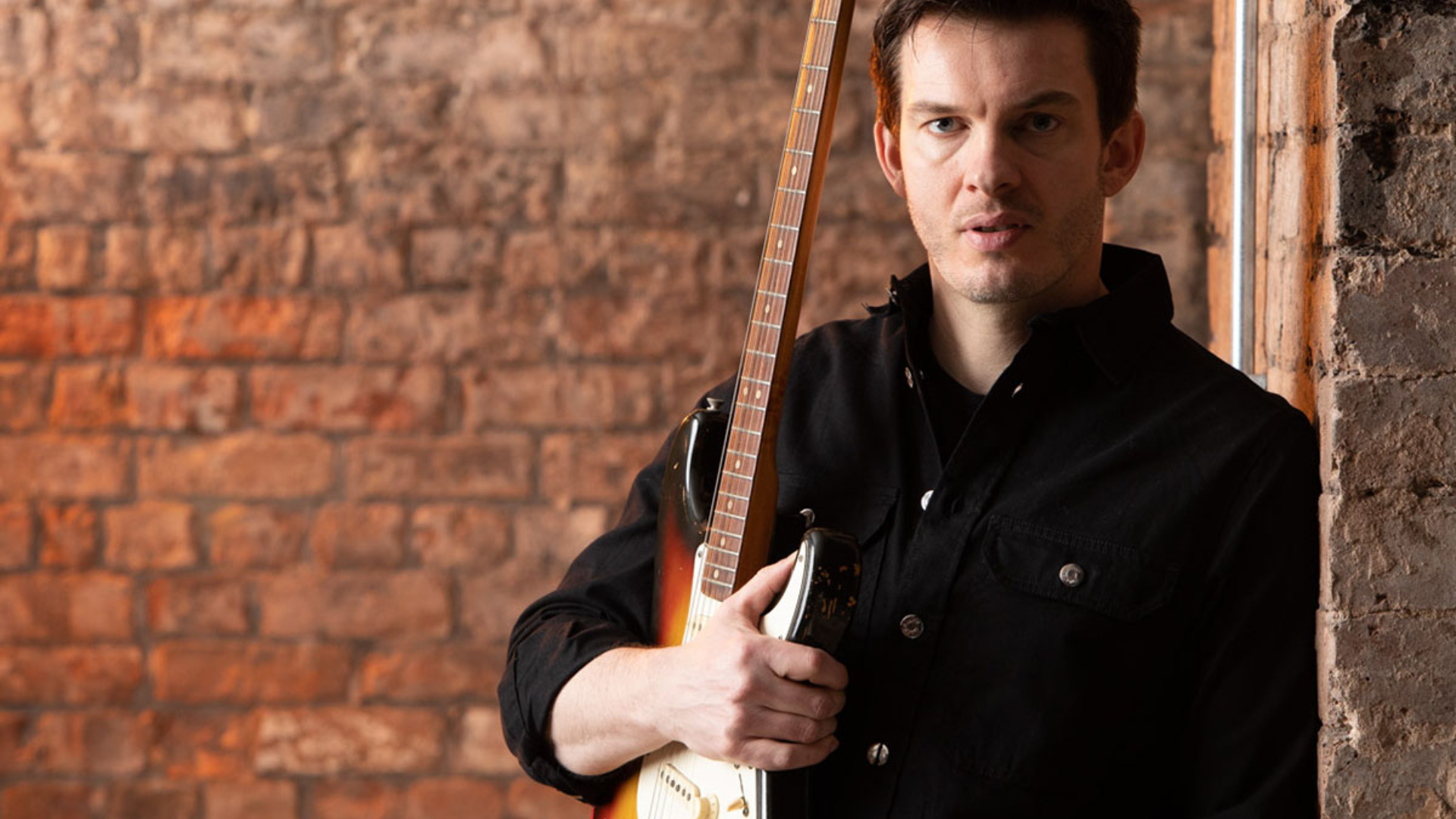 Dan Machin leans against a brick wall holding his Fender guitar